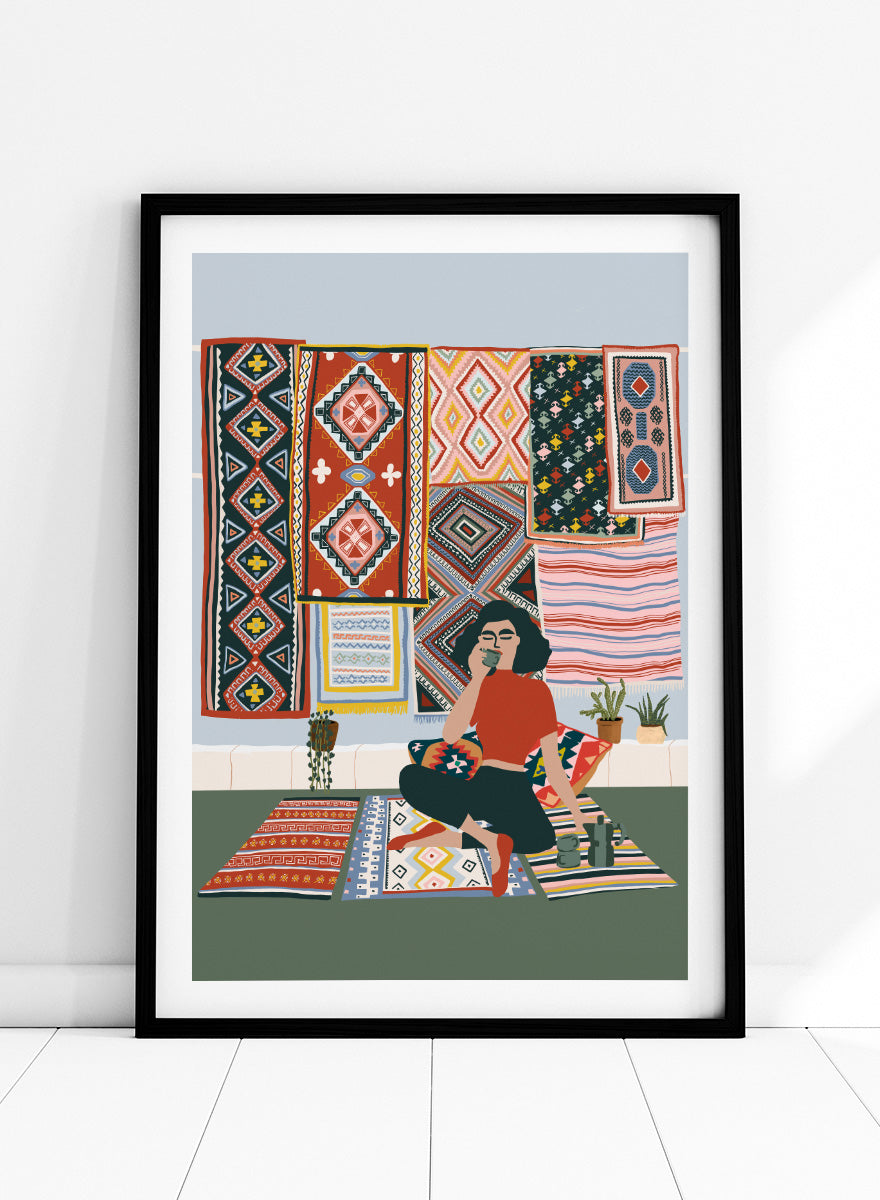 A framed art print featuring a stylized illustration of a woman sitting cross-legged on a patterned rug, surrounded by colorful geometric textiles hanging on a wall. Bohemian Coffee Morning by Ceyda Alasar interior art print featuring patterned rugs, floor cushions, potted plants, coffee, and colorful collected decor.