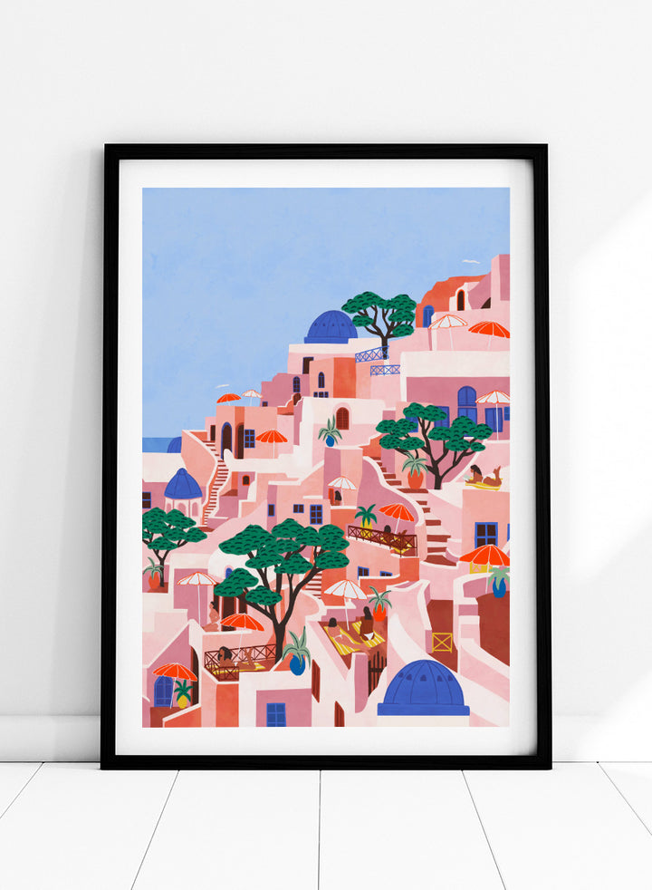 A stylized art print depicting a hillside Mediterranean village with pink and peach buildings, blue domes, green trees, and a light blue sky. Santorini Summer Dream by Ceyda Alasar art print featuring blue domes, terraced Mediterranean architecture, orange umbrellas, and sunny Greek island charm.