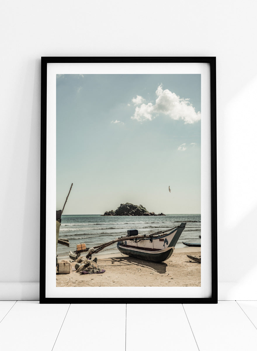 A framed photographic print of a coastal beach scene with a small boat on the sandy shore, the sea and distant rocky island, and a pale blue sky with clouds. Seaside Serenity by Shot by Clint coastal photography print featuring a fishing boat, sandy shore, ocean waves, distant island, and weathered seaside details.
