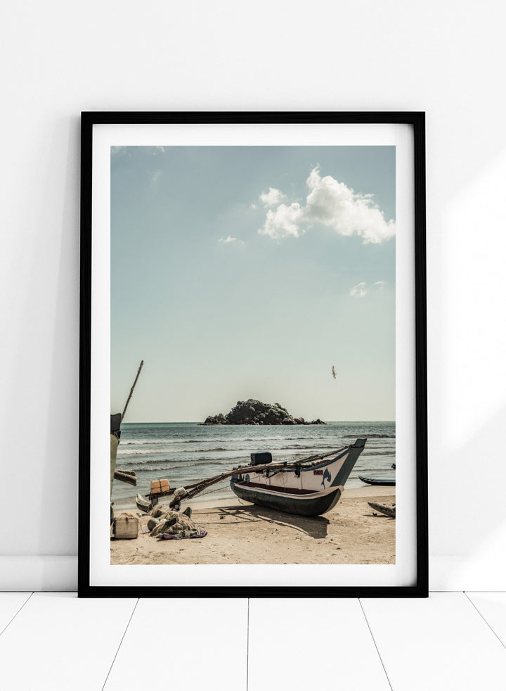 A framed photographic print of a coastal beach scene with a small boat on the sandy shore, the sea and distant rocky island, and a pale blue sky with clouds. Seaside Serenity by Shot by Clint coastal photography print featuring a fishing boat, sandy shore, ocean waves, distant island, and weathered seaside details.