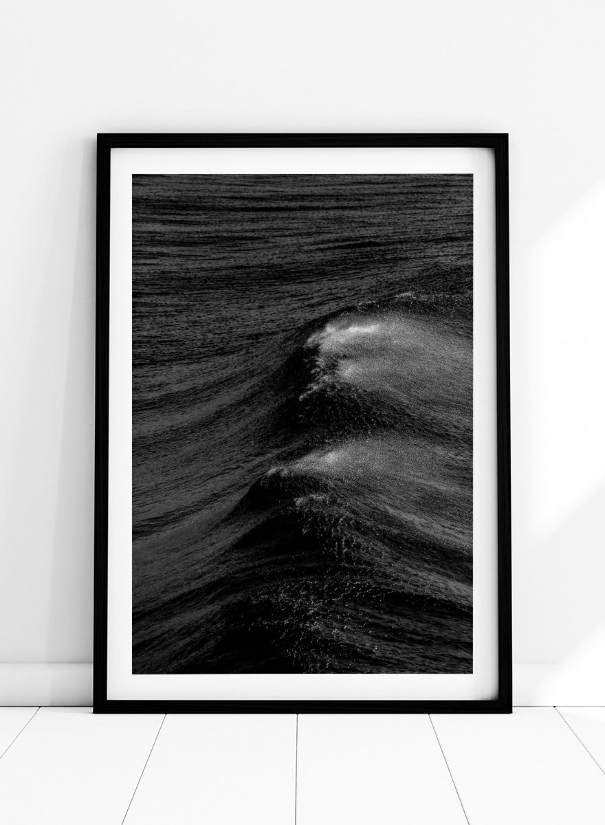 Black and white fine art photographic print of ocean waves, featuring a dramatic wave crest with white foam against dark textured water. Night Wave Cascade by Shot by Clint black-and-white ocean photography print featuring dark rolling waves, sea spray, and dramatic natural movement.