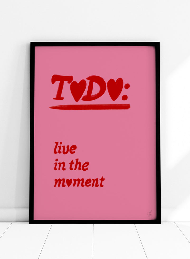 Pink poster with red text 'TODO: live in the moment' on a pink background. Typography wall art reading “live in the moment” with red hearts on pink background by Anne-Marie Volfova. Officially licensed motivational art print.