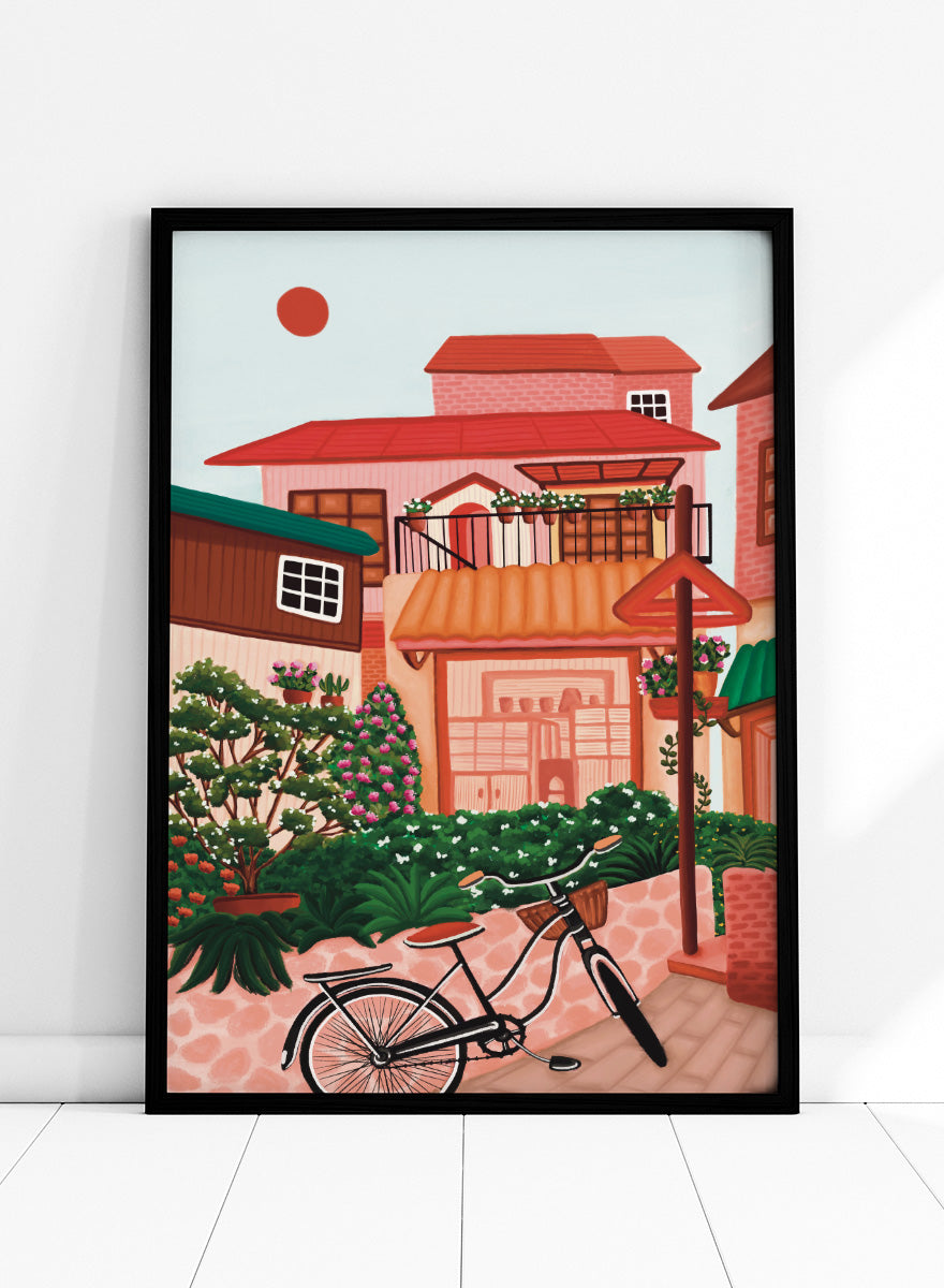 A colorful illustrated street scene with pink and peach buildings, green plants, flowers, a bicycle in the foreground, and a pale sky with a red sun. Shop Charming Village Street by Nidhi Kachhadiya, a colorful village wall art print featuring a bicycle, flower-filled homes, terracotta roofs, and warm pink and green tones.