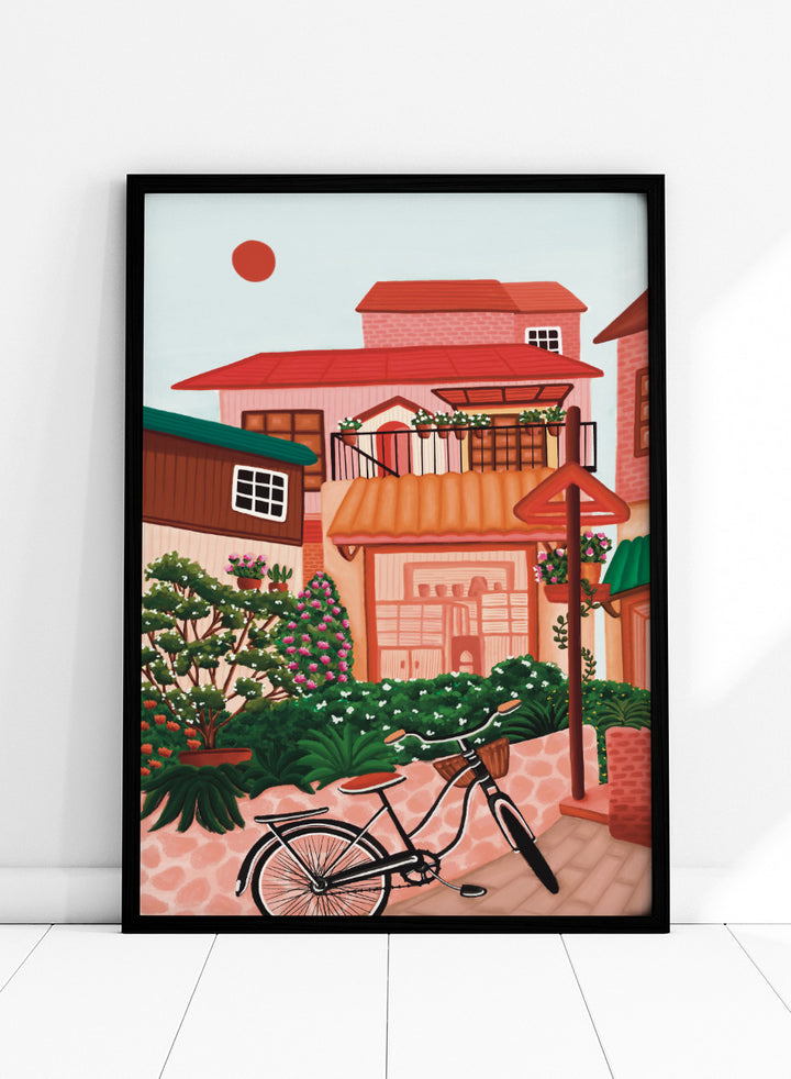 A colorful illustrated street scene with pink and peach buildings, green plants, flowers, a bicycle in the foreground, and a pale sky with a red sun. Shop Charming Village Street by Nidhi Kachhadiya, a colorful village wall art print featuring a bicycle, flower-filled homes, terracotta roofs, and warm pink and green tones.