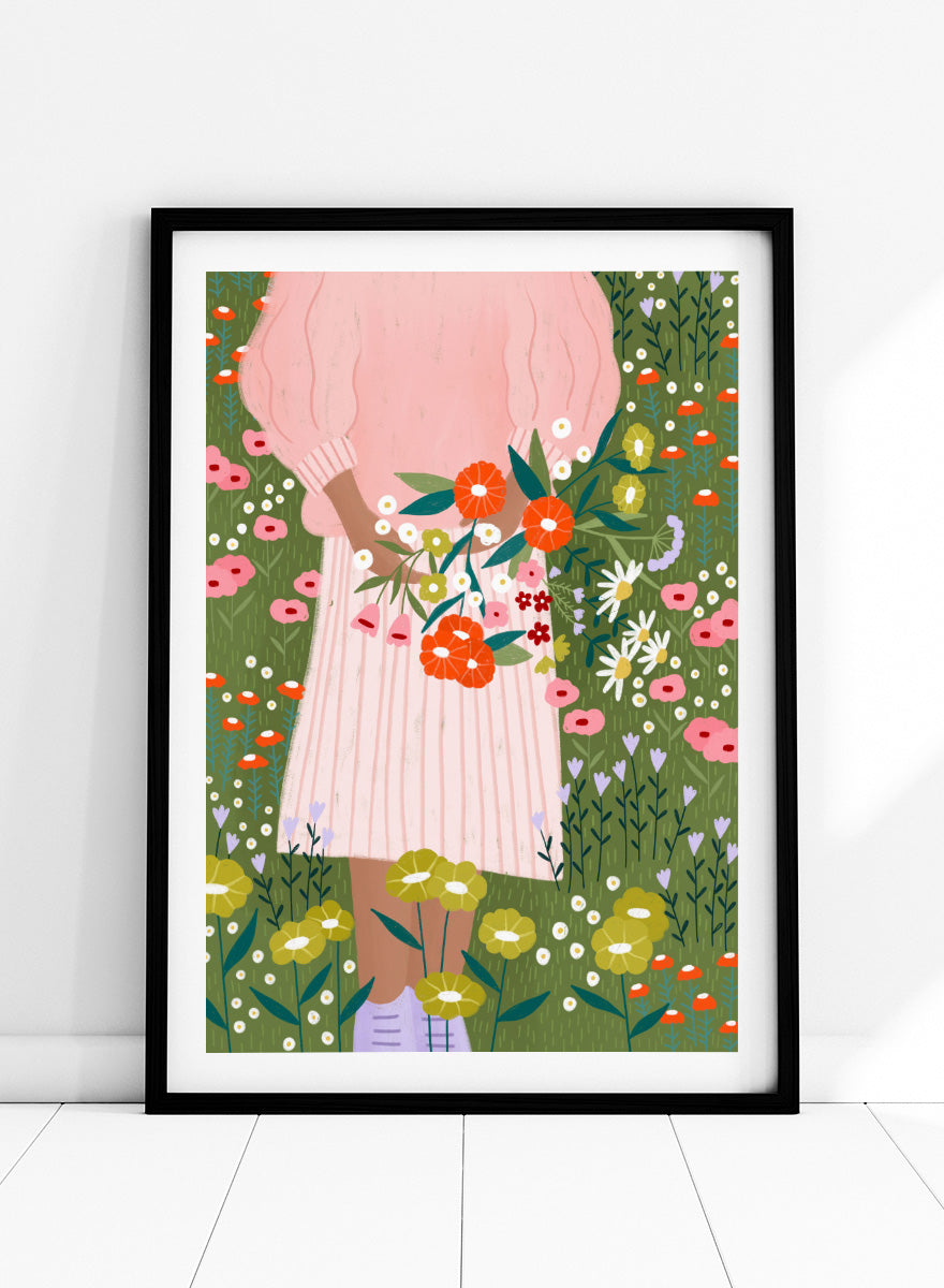 A framed art print featuring a stylized illustration of a person in a pink striped dress holding a bouquet of orange, red, and white flowers in a green field of wildflowers. Secret Garden Bouquet by Bea Müller floral art print featuring a woman gathering wildflowers in a colorful garden filled with poppies, daisies, and greenery.