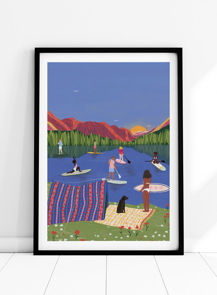 A framed art print featuring a colorful, stylized illustration of people paddleboarding and relaxing on a lake with mountains in the background. Paddleboard Lake Life by Ceyda Alasar outdoor art print featuring paddleboarders, mountain scenery, wildflowers, picnic blankets, and a lakeside dog.