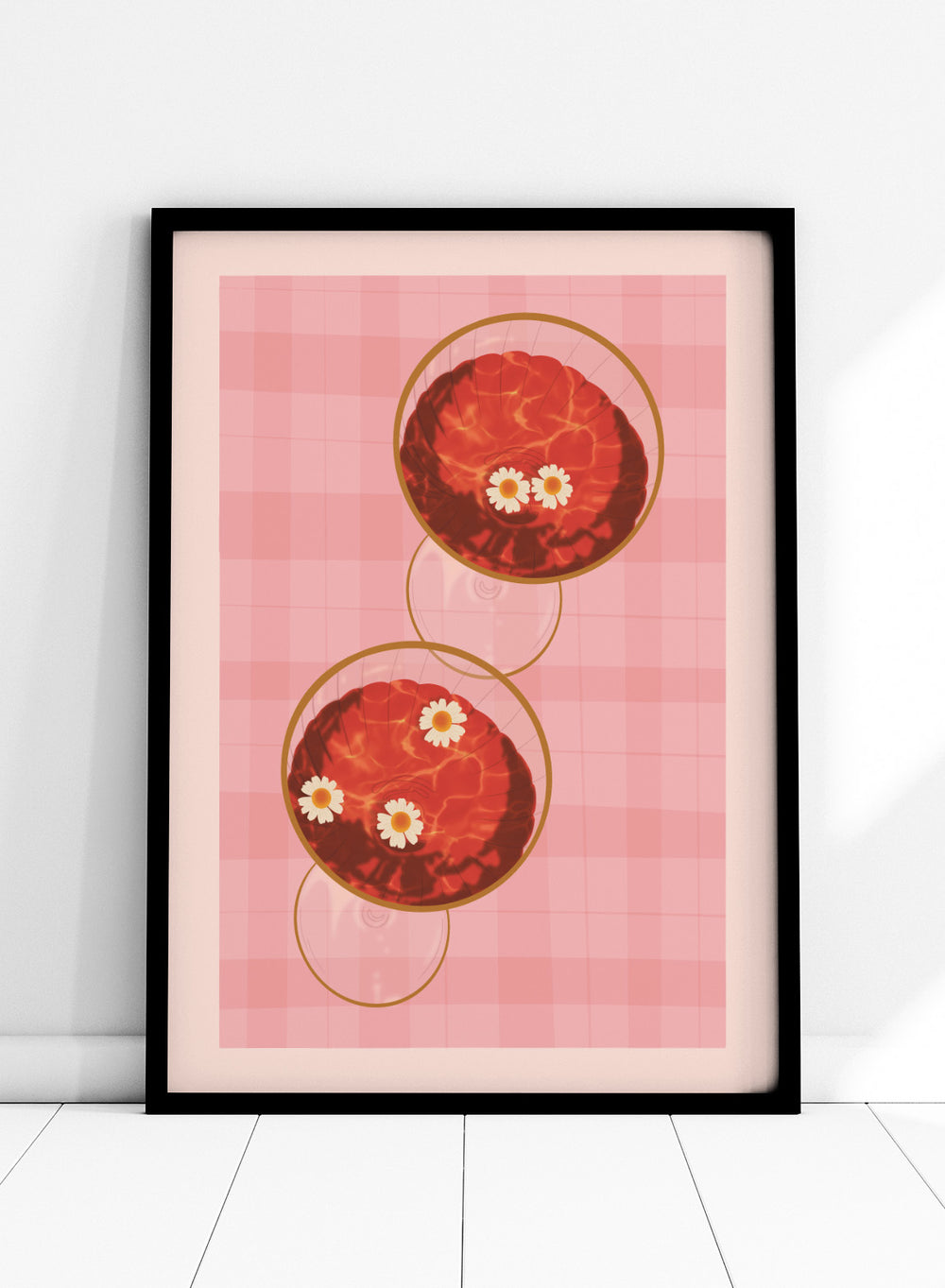 A stylized art print featuring two overlapping pink and red plaid glasses with white daisy flowers, framed by a black border on a light pink background. Pink Drinks With Daisies by Lisa Solomoniuk premium framed fine art print featuring red coupe glasses, floating daisies, and pink gingham.