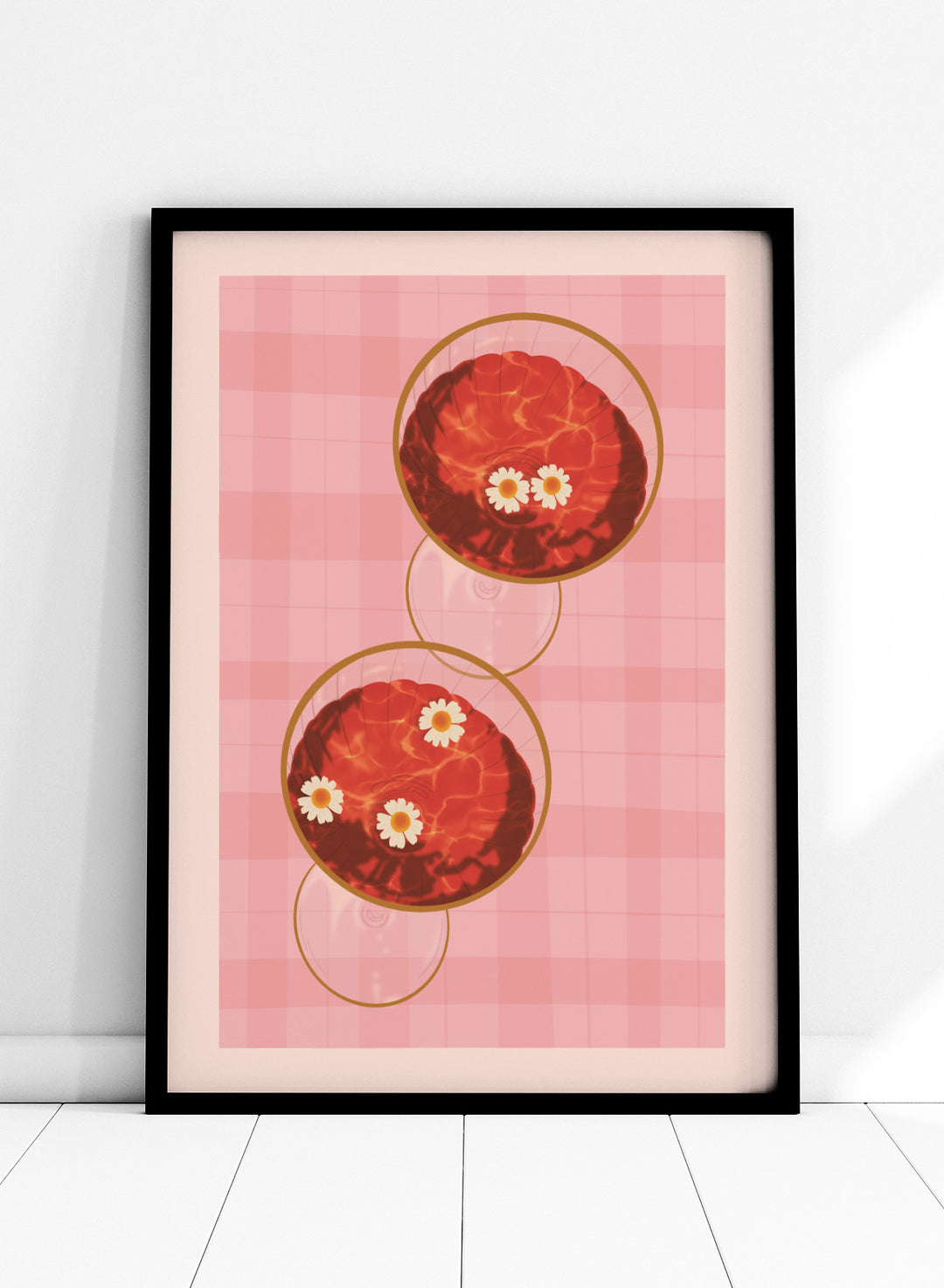 A stylized art print featuring two overlapping pink and red plaid glasses with white daisy flowers, framed by a black border on a light pink background. Pink Drinks With Daisies by Lisa Solomoniuk premium framed fine art print featuring red coupe glasses, floating daisies, and pink gingham.