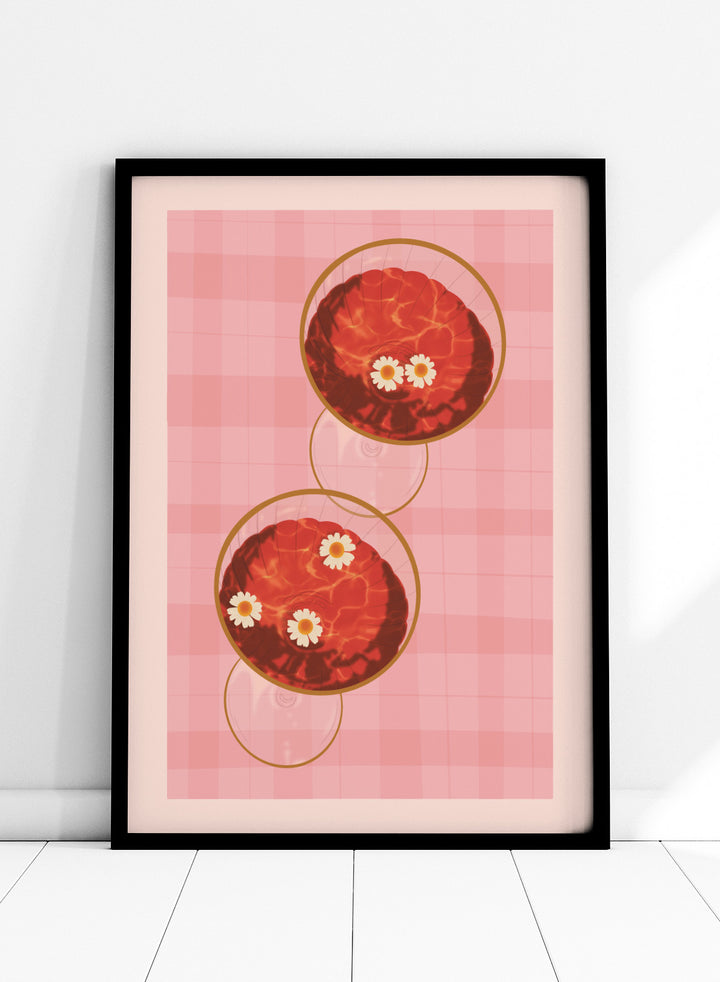 A stylized art print featuring two overlapping pink and red plaid glasses with white daisy flowers, framed by a black border on a light pink background. Pink Drinks With Daisies by Lisa Solomoniuk premium framed fine art print featuring red coupe glasses, floating daisies, and pink gingham.