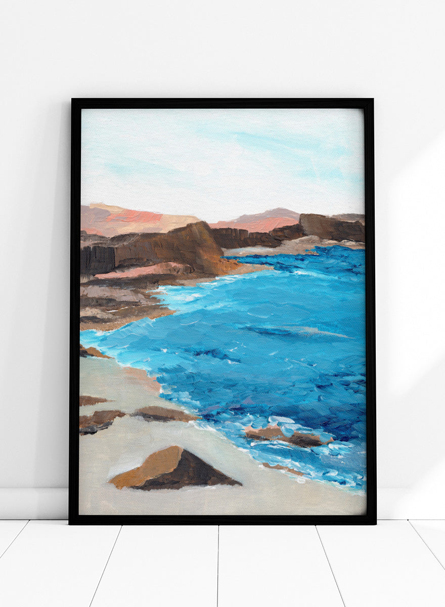 A framed landscape painting depicting a coastal scene with bright blue ocean water, sandy shore, dark rocky cliffs, and distant hills under a pale sky. Shop Rocky Coastal Waters by Nikita Jariwala, a coastal wall art print poster with blue ocean water, rocky cliffs, and a peaceful seaside view.