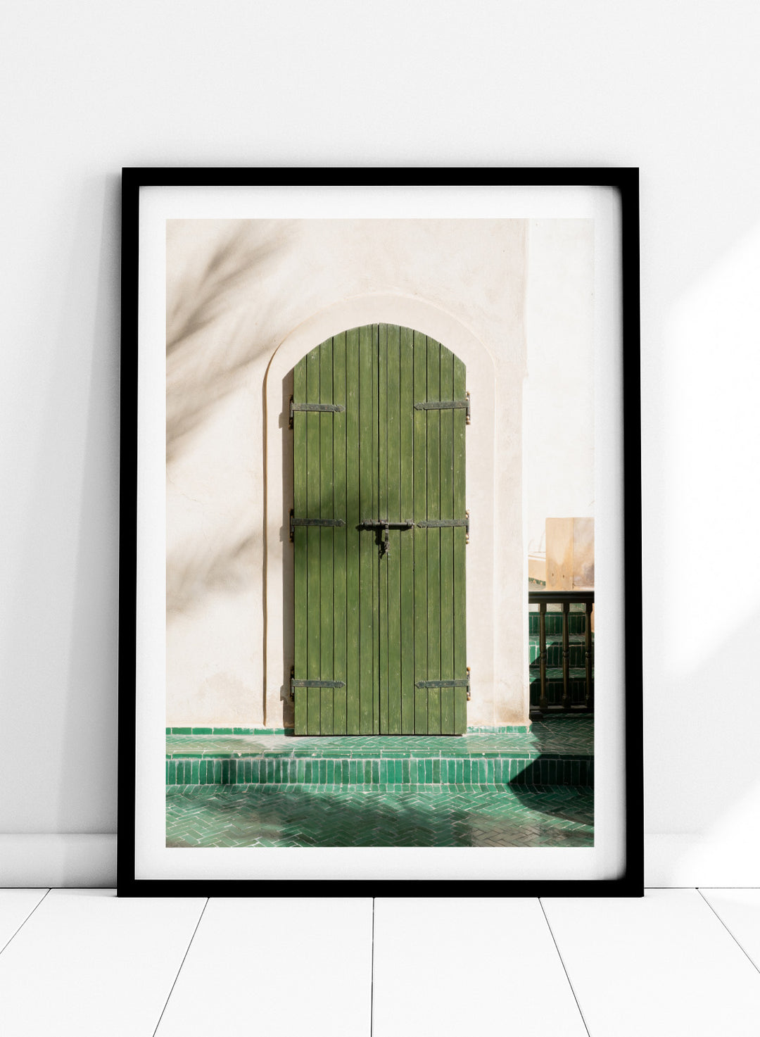 Green wooden door with metal latch on a white wall next to a pool. Moroccan Green Door by Henrike Schenk is an architectural photography print featuring green tilework, a traditional doorway, and Moroccan design details.