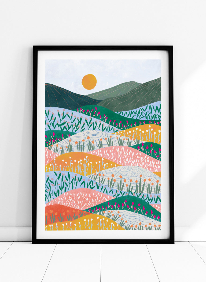 A framed art print featuring a stylized landscape illustration with rolling hills, wildflowers, mountains, and a sun in pastel and bright colors. Colorful Flower Hills by Ceyda Alasar botanical landscape art print featuring layered wildflower fields, rolling hills, mountains, and a golden sun.