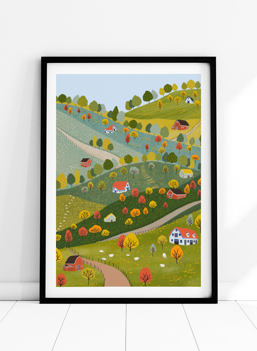 A framed art print featuring a whimsical, stylized illustration of a rolling green and yellow countryside landscape with red houses, colorful autumn trees, winding roads, and a pale blue sky. Vibrant Autumn Village Hills by Ceyda Alasar art print with rolling countryside, colorful fall trees, farmhouses, barns, sheep, and winding roads.
