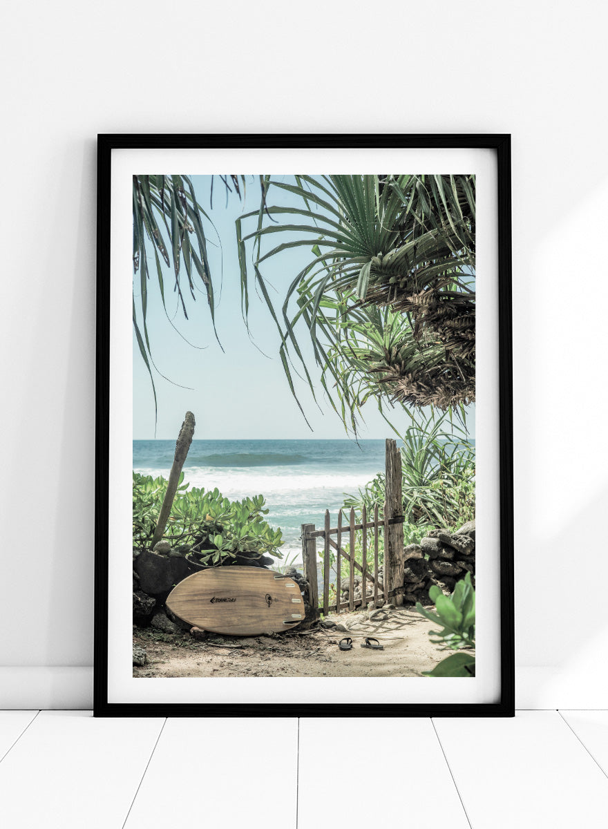 A framed beach scene art print featuring a wooden surfboard leaning against a rustic gate, with palm leaves framing a view of the ocean under a light blue sky. Coastal Serenity by Shot by Clint coastal art print featuring a surfboard, tropical plants, rustic gate, sandy path, and ocean waves.