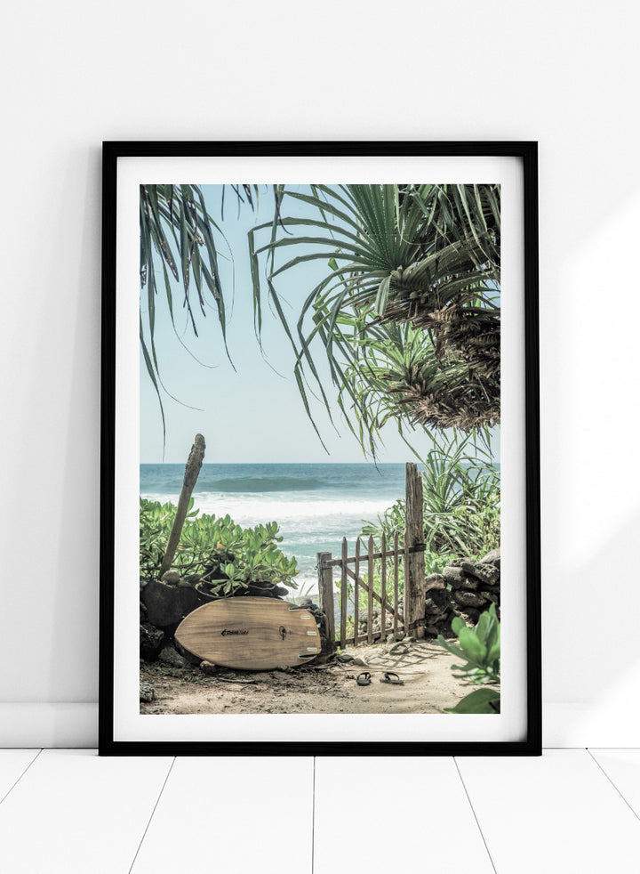 A framed beach scene art print featuring a wooden surfboard leaning against a rustic gate, with palm leaves framing a view of the ocean under a light blue sky. Coastal Serenity by Shot by Clint coastal art print featuring a surfboard, tropical plants, rustic gate, sandy path, and ocean waves.
