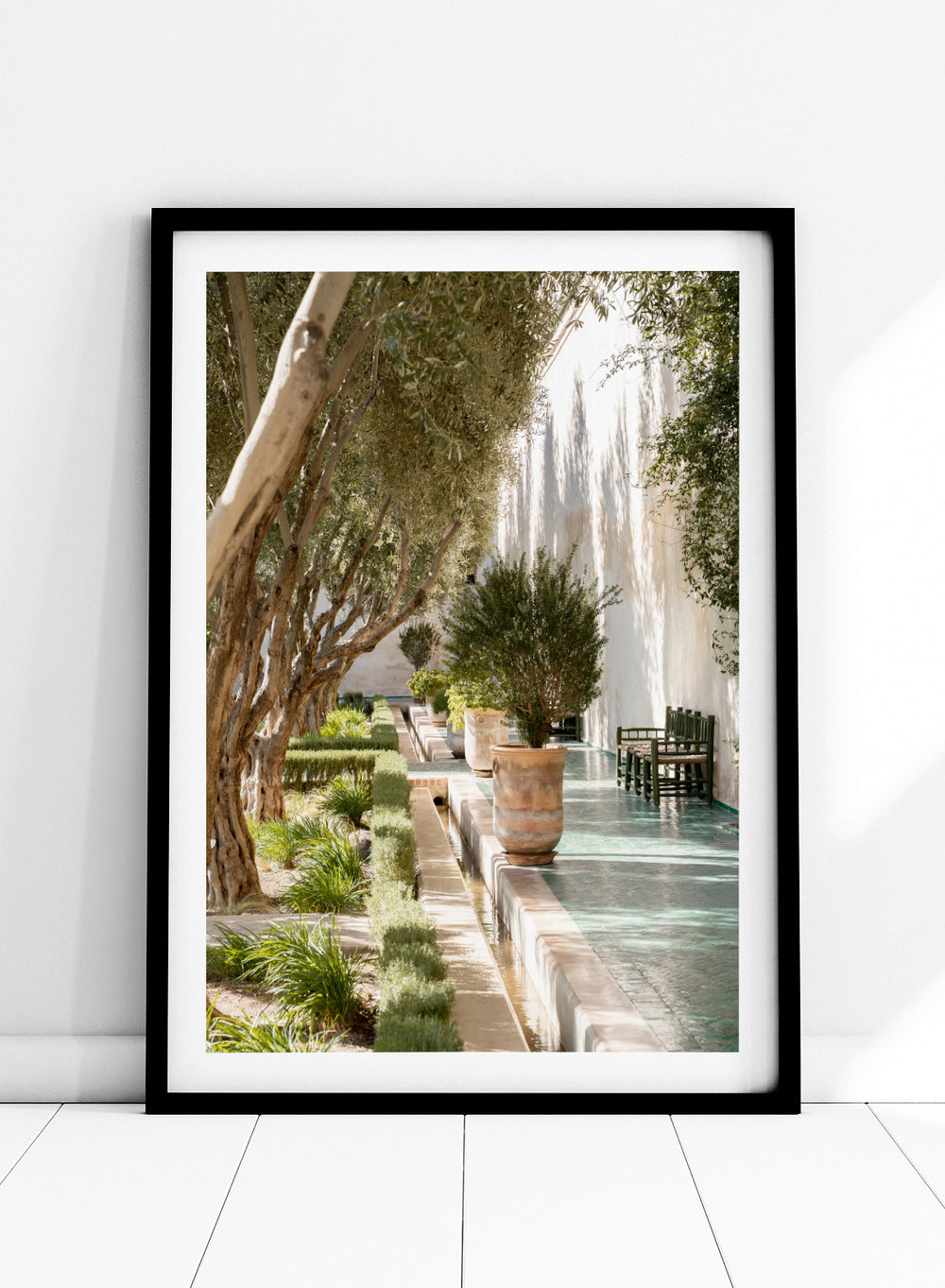 Garden pathway with trees, plants, and a building in the background. Mediterranean Garden Courtyard by Henrike Schenk is a travel photography print featuring olive trees, terracotta pots, tiled walkways, and a Moroccan garden.