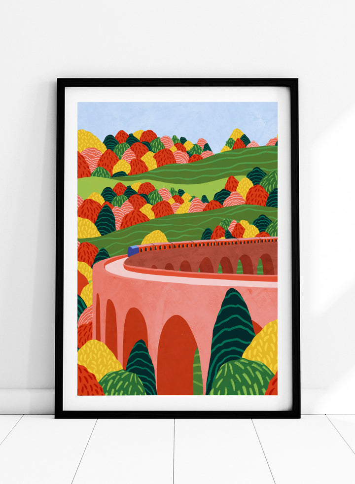 A framed art print featuring a stylized landscape illustration with rolling green hills, colorful patterned trees, and a pink arch bridge under a light blue sky. Winding Autumn Viaduct by Ceyda Alasar travel art print featuring a colorful train, arched bridge, rolling hills, and bold autumn foliage.