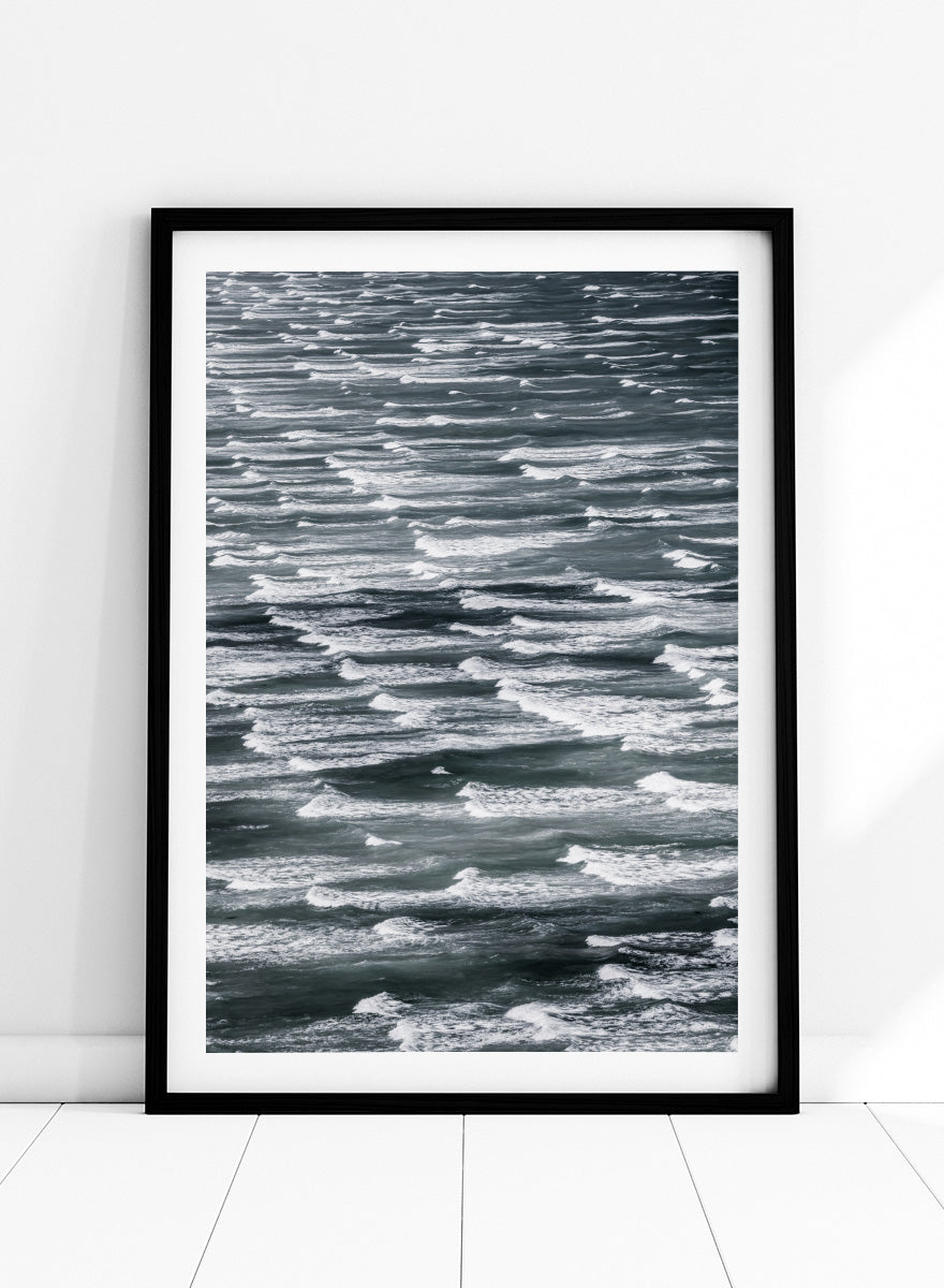 A framed black and white photographic print depicting a close-up view of ocean waves with white foam crests against dark water. Waves in Rhythm by Shot by Clint ocean photography print featuring layered blue-grey waves, white foam, and powerful natural movement across the sea.