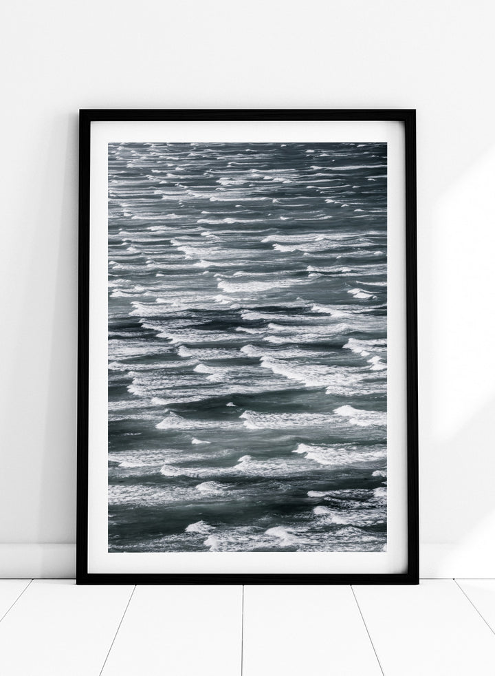 A framed black and white photographic print depicting a close-up view of ocean waves with white foam crests against dark water. Waves in Rhythm by Shot by Clint ocean photography print featuring layered blue-grey waves, white foam, and powerful natural movement across the sea.