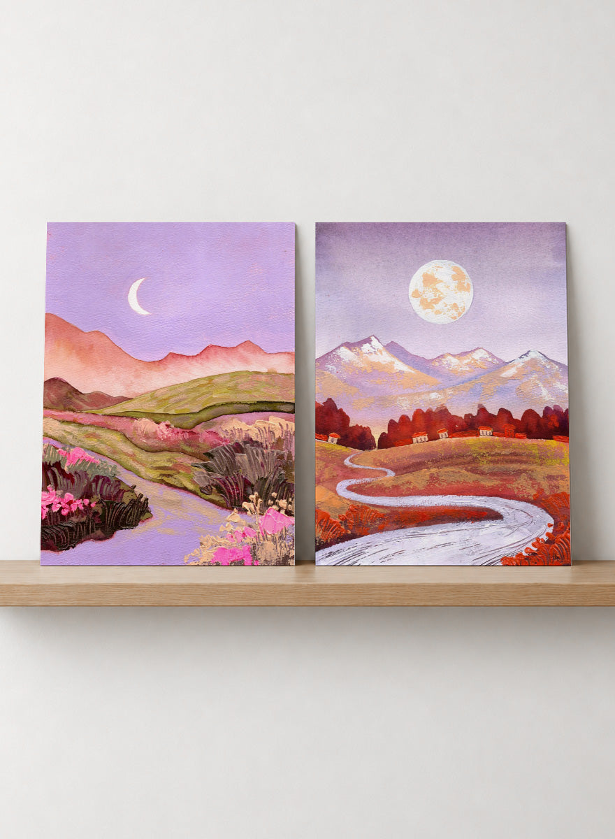 Two framed landscape art prints displayed side by side on a black shelf against a light beige wall. The left print shows a purple-toned valley with a crescent moon, pink flowers, and distant hills. Shop Moonlit Landscapes by Ekaterina Prisich, a set of 2 landscape wall art prints posters featuring moonlit mountains, winding paths, and purple tones.