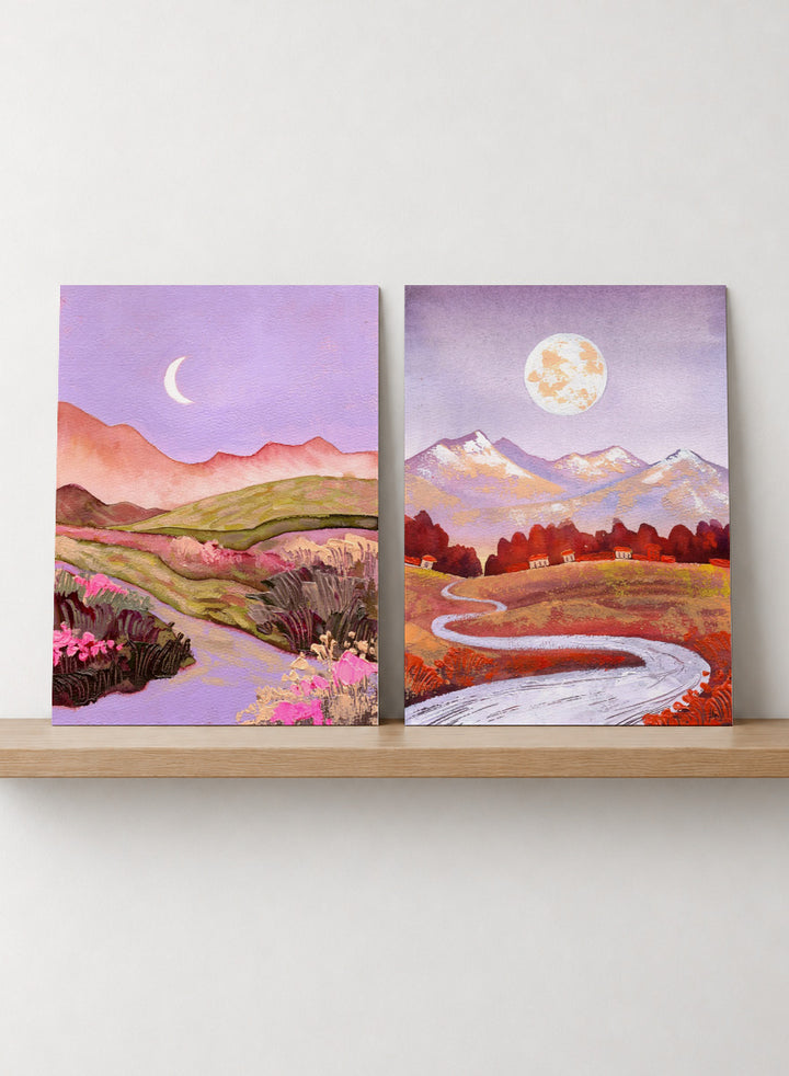 Two framed landscape art prints displayed side by side on a black shelf against a light beige wall. The left print shows a purple-toned valley with a crescent moon, pink flowers, and distant hills. Shop Moonlit Landscapes by Ekaterina Prisich, a set of 2 landscape wall art prints posters featuring moonlit mountains, winding paths, and purple tones.