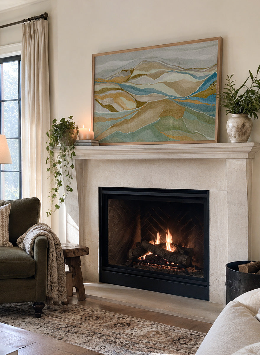 Framed abstract landscape wall art featuring layered hills and winding blue river-like lines in beige, tan, muted green, and blue tones, shown against a light neutral background. Shop Abstract Mountain Serenity by Nikita Jariwala, a modern mountain wall art print featuring layered abstract hills, earthy tones, and soft blue and sage accents.