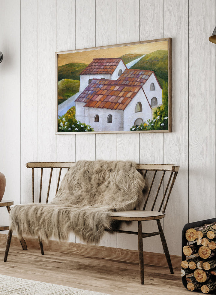 A framed landscape painting depicting two white buildings with terracotta tiled roofs nestled in green hills, with a winding light blue road and a warm yellow sky. Shop Tuscan Hillside Village by Angeles M. Pomata, a warm countryside landscape art print featuring white houses, terracotta roofs, green hills, and wildflowers.