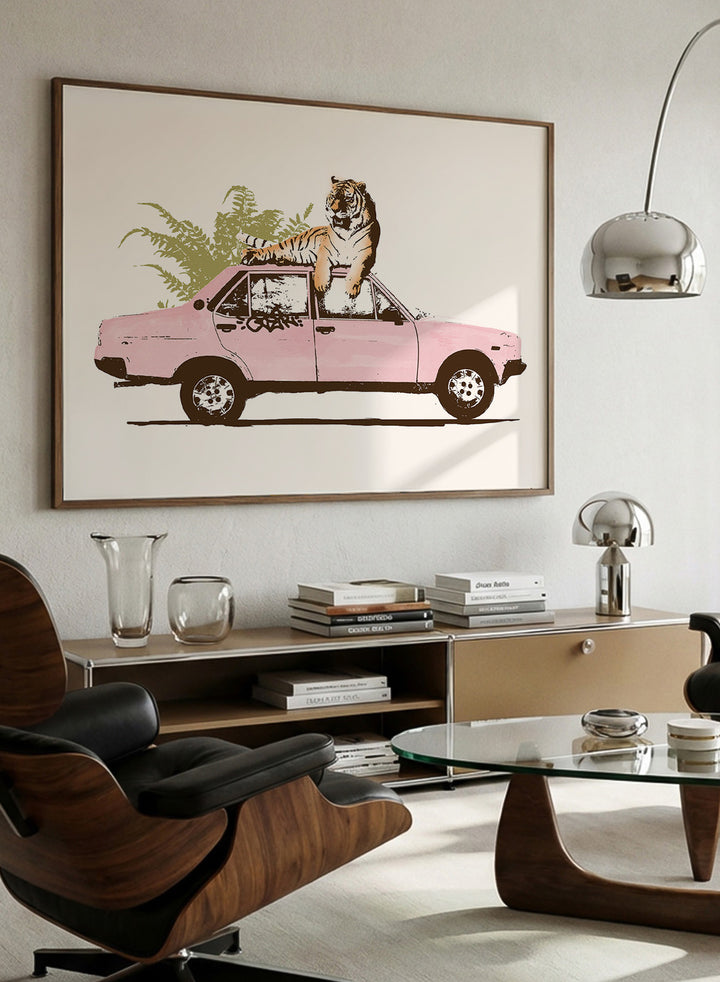 Framed artwork of a tiger in a pink car with greenery on a white background. Tiger On Pink Car by Florent Bodart is a surreal animal art print with a tiger on a vintage car, a modern wall poster with playful, bold, and imaginative design.
