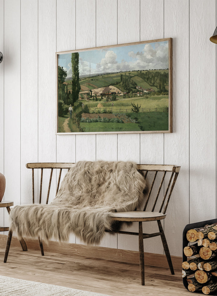 A framed landscape painting depicting a green countryside scene with rolling hills, trees, a path, small buildings, and a cloudy sky. Landscape at Les Pâtis, Pontoise by Camille Pissarro is a vintage French landscape print featuring green fields, farm workers, rolling hills, and a summer sky.
