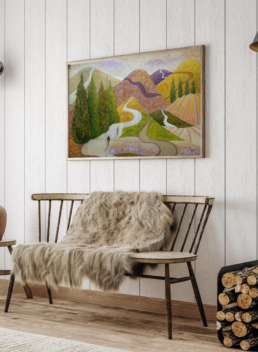 A framed landscape painting featuring stylized mountains, evergreen trees, winding paths, and small human figures, shown against a light neutral background. Shop Peaceful Journey Through Hills by Angeles M. Pomata, an autumn landscape art print featuring winding roads, cypress trees, rolling hills, and two walking figures.