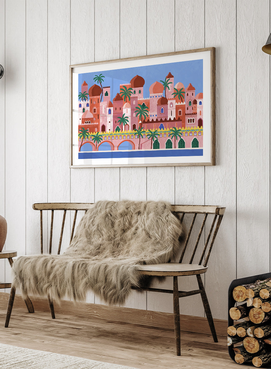 A framed art print featuring a stylized illustration of a coastal city with pink and peach buildings, green palm trees, and a blue sky. Arabian Fantasy City by Ceyda Alasar travel art print featuring ornate domes, arches, palm trees, patterned facades, and colorful desert architecture.