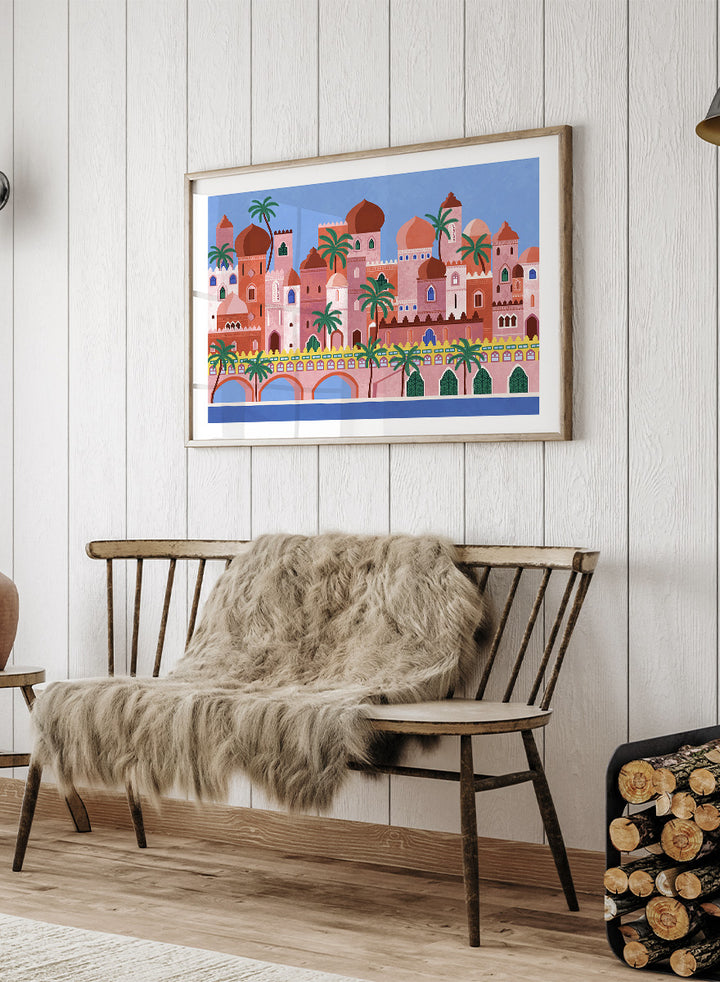 A framed art print featuring a stylized illustration of a coastal city with pink and peach buildings, green palm trees, and a blue sky. Arabian Fantasy City by Ceyda Alasar travel art print featuring ornate domes, arches, palm trees, patterned facades, and colorful desert architecture.