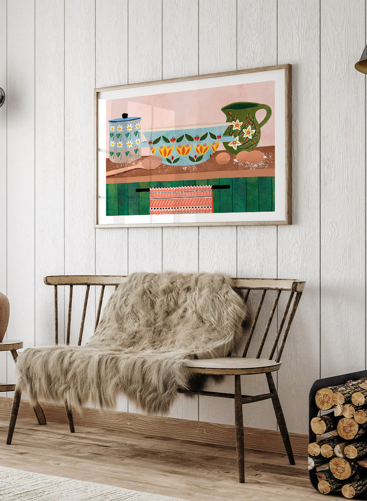 Framed art print featuring a whimsical still life illustration of kitchenware, including a floral-patterned bowl, a green pitcher, eggs, and a patterned towel. Charming Folk Kitchen by Ceyda Alasar kitchen art print featuring painted pottery, floral patterns, wooden spoons, eggs, and colorful folk-inspired decor.