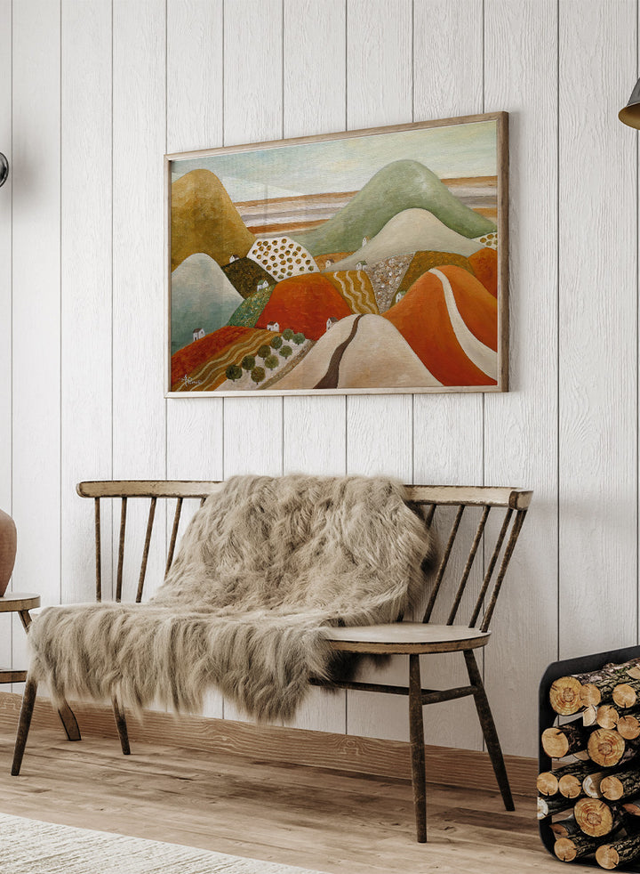 A framed landscape painting featuring stylized rolling hills and fields in earthy tones of orange, green, beige, and brown. Shop Colorful Hillside Village by Angeles M. Pomata, an autumn landscape art print featuring rolling hills, tiny homes, winding roads, and warm earthy tones.