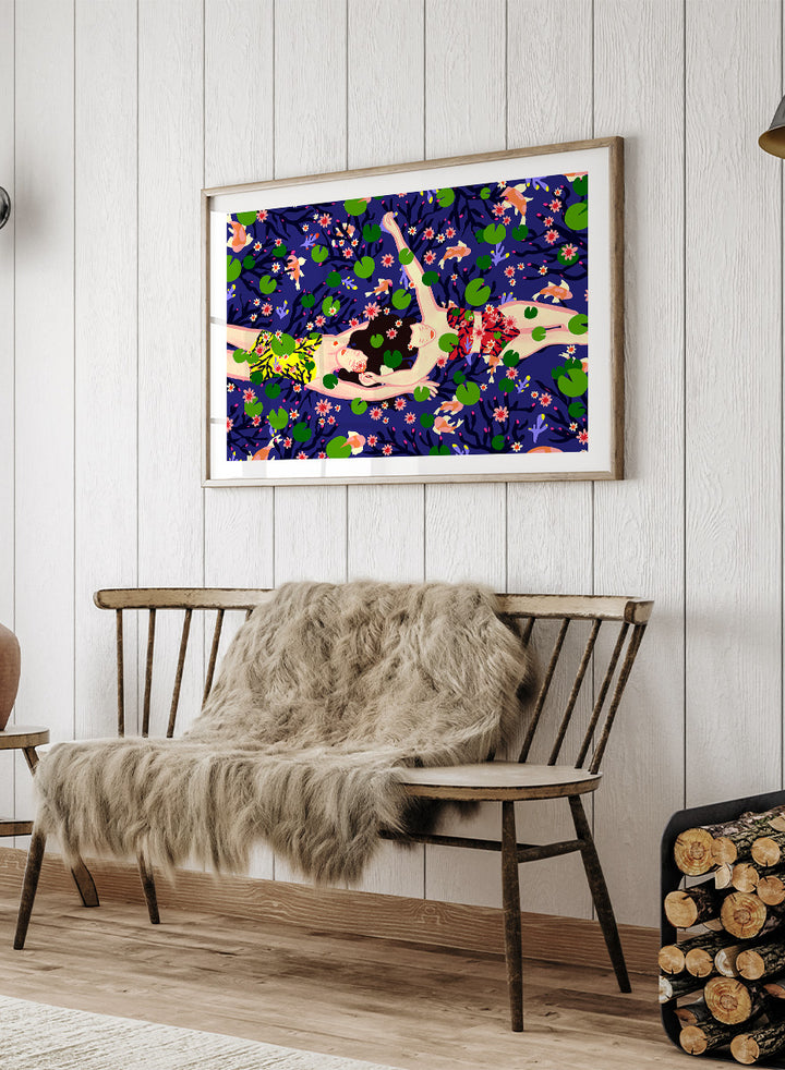 A framed art print featuring a stylized illustration of a woman lying on a dark blue background covered with green lily pads and pink flowers. Serene Night Float by Ceyda Alasar friendship art print featuring three women floating among lily pads, flowers, and fish in a deep blue pond.