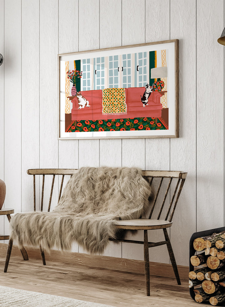 A framed art print depicting a cozy living room scene with a coral-pink sofa, two cats, a patterned throw blanket, and a floral rug. Playful Cats Living Room by Ceyda Alasar cat art print featuring two mischievous cats, a coral sofa, floral rug, patterned throw, and colorful decor.