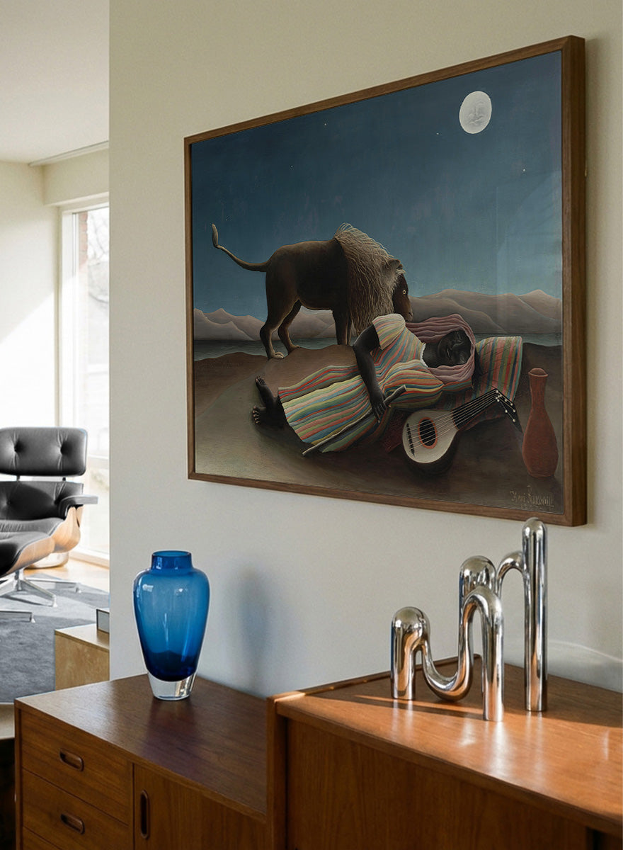 A framed surreal art print depicting a lion standing over a sleeping person wrapped in a striped blanket, with a guitar, vase, and full moon in a night sky. The Sleeping Gypsy by Henri Rousseau is a vintage art print featuring a sleeping traveler, lion, moonlit desert, striped robe, mandolin, and deep blue night sky.