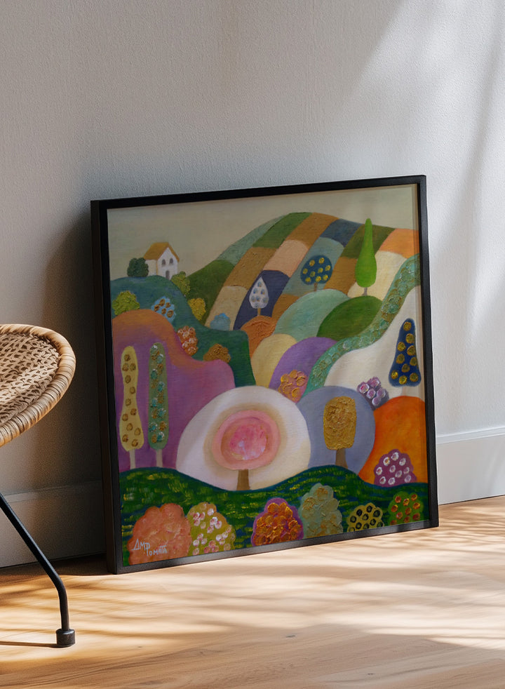 A colorful abstract landscape painting in a black frame, featuring stylized hills, trees, flowers, and a small house. The artwork has a playful, patchwork-like design with multiple vibrant colors including green, purple, orange, pink, blue, and yellow, set against a light beige wall. Shop Whimsical Hillside Village by Ángeles M. Pomata, a colorful landscape art print with rolling hills, patterned trees, and a dreamy village feel.