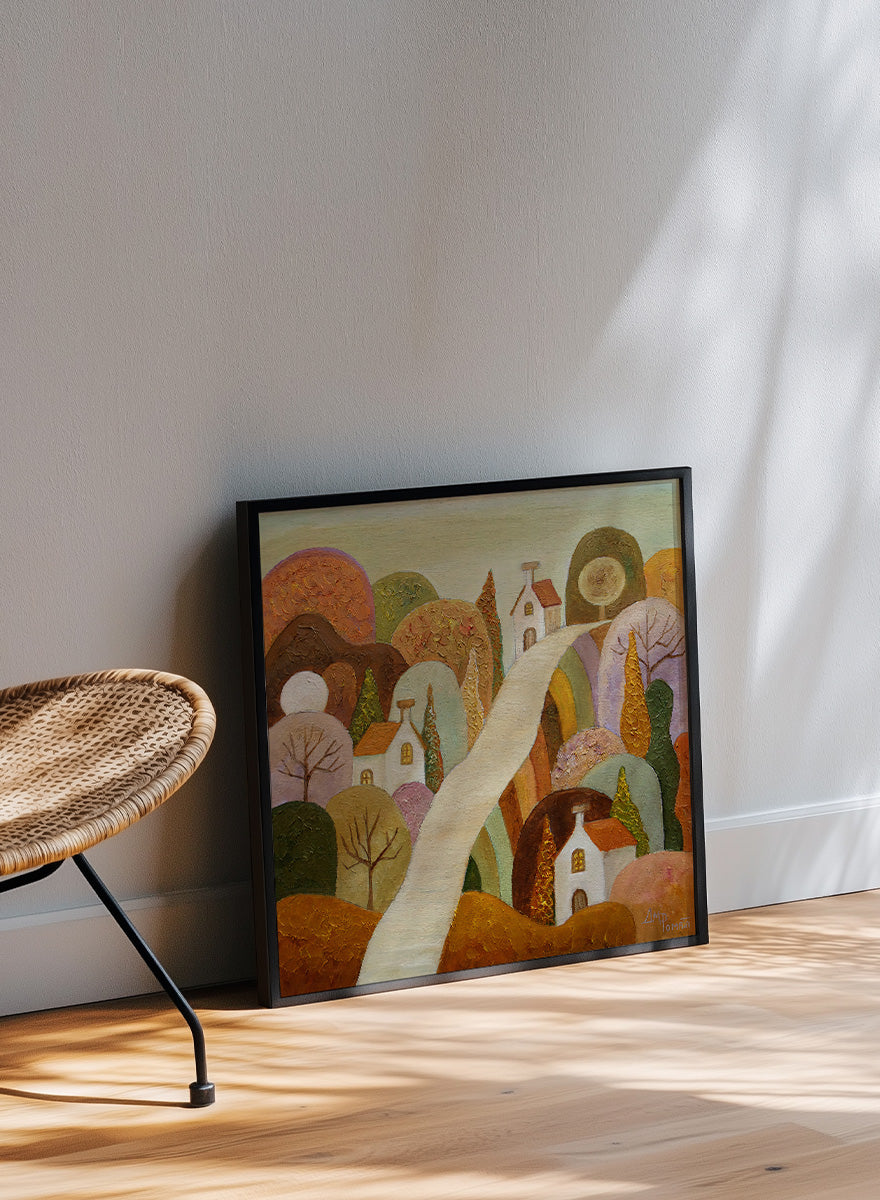 A framed landscape painting featuring stylized white houses, a winding cream path, and rolling hills in earthy tones of brown, orange, green, and purple. Shop Whimsical Village Path by Angeles M. Pomata, an autumn landscape art print featuring a winding road, white village houses, rounded hills, and warm earthy tones.