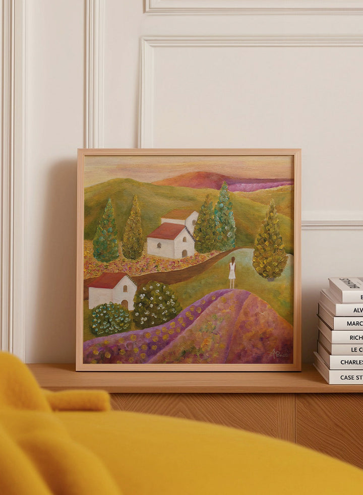 A framed landscape painting featuring rolling green hills, white houses with red roofs, tall conical trees, and a figure standing on a purple field. Shop Woman in Lavender Fields by Angeles M. Pomata, a colorful landscape and figurative art print featuring lavender fields, white houses, cypress trees, and rolling hills.