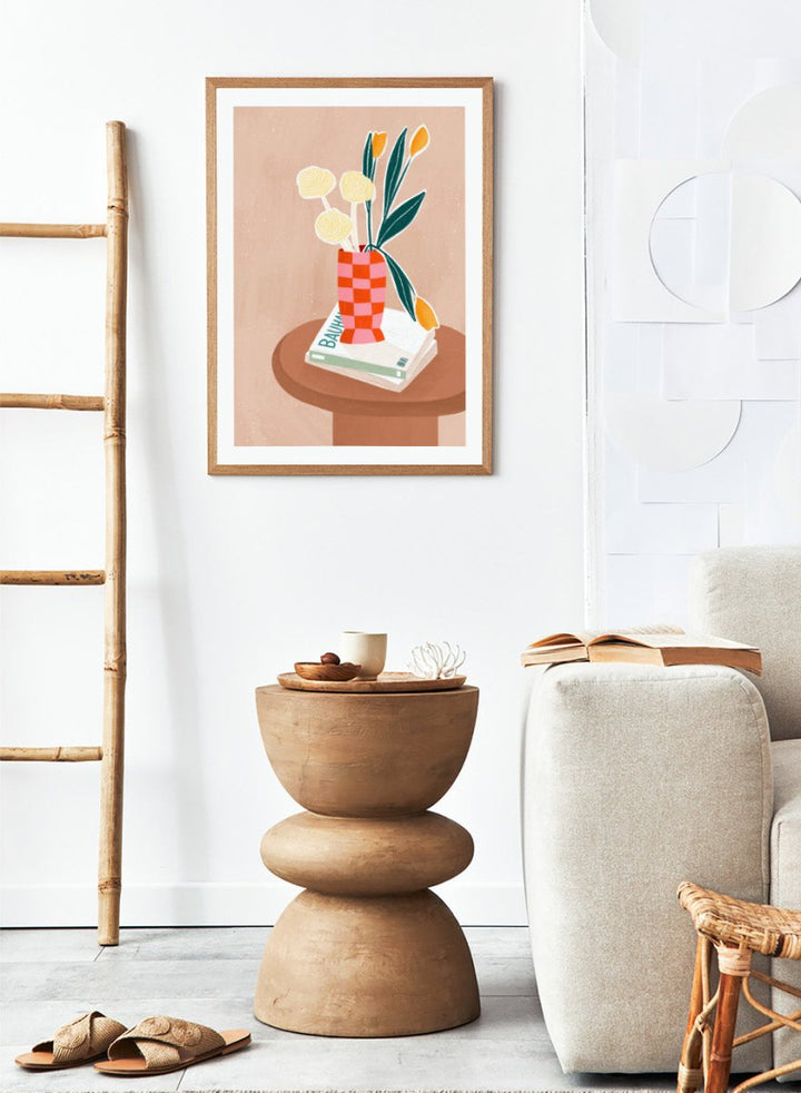 A framed minimalist art print featuring a stylized illustration of a pink and orange checkered vase filled with yellow and orange tulips, resting on a brown round table next to a white book. Colorful Tabletop Flowers by Bea Müller premium framed fine art print featuring yellow tulips and cream blooms in a red-and-pink checkered vase.