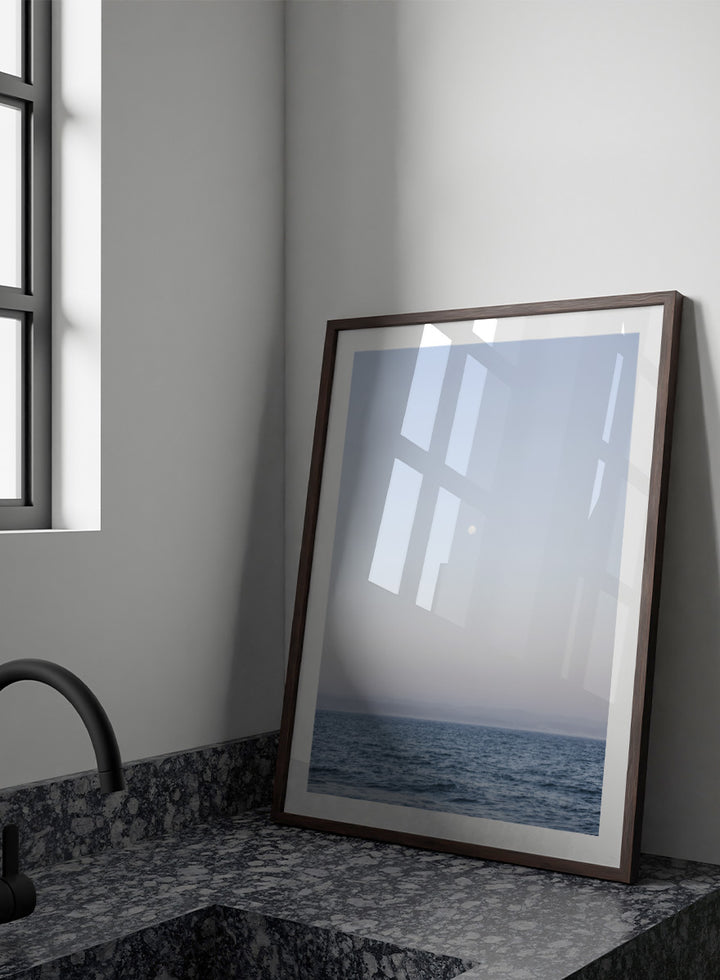 A minimalist framed art print depicting a calm blue ocean under a pale sky with a small visible moon. Moonlit Horizon by Shot by Clint coastal photography print featuring a pale moon, distant shoreline, open blue water, and soft lavender evening tones.