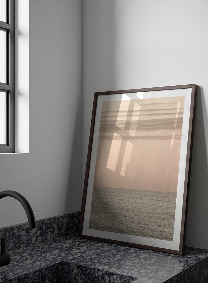 A minimalist framed art print depicting a calm seascape at dusk or dawn, with a soft peach and beige sky meeting a muted gray-blue ocean horizon. Serene Horizons by Shot by Clint features a soft peach sky and calm ocean horizon in a minimal coastal photograph with airy neutral tones.
