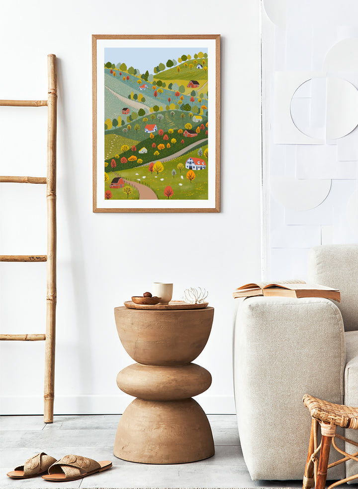 A framed art print featuring a whimsical, stylized illustration of a rolling green and yellow countryside landscape with red houses, colorful autumn trees, winding roads, and a pale blue sky. Vibrant Autumn Village Hills by Ceyda Alasar art print with rolling countryside, colorful fall trees, farmhouses, barns, sheep, and winding roads.