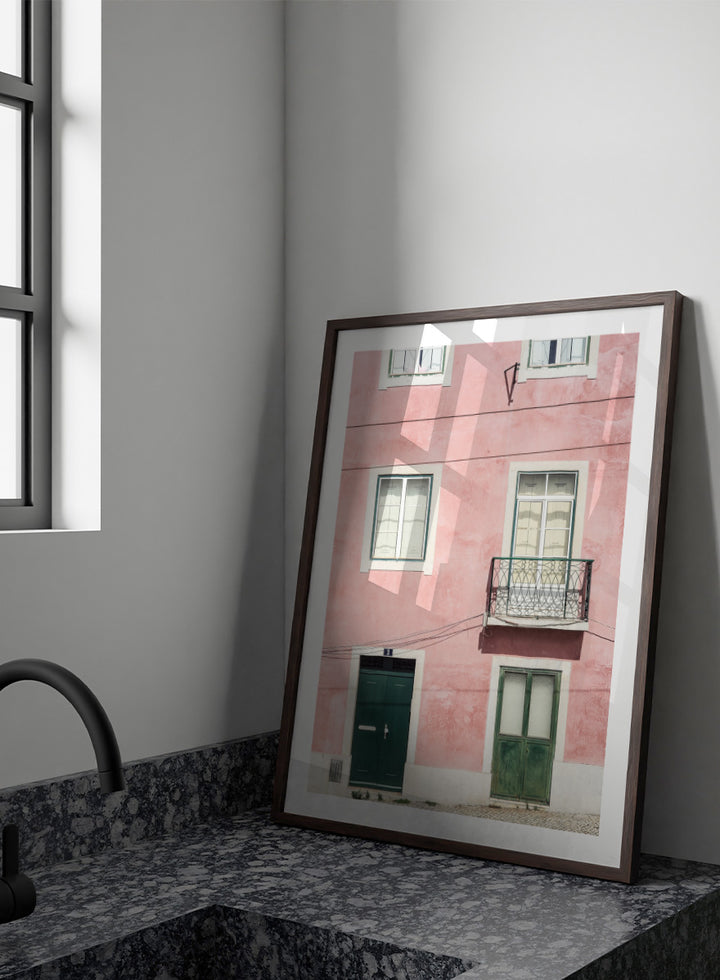 A framed art print featuring a photograph of a pink building facade with green doors, white-framed windows, and a small black metal balcony. Pink Façade Harmony by Shot by Clint architectural photography print featuring a pink exterior, green doors, white windows, and an iron balcony.