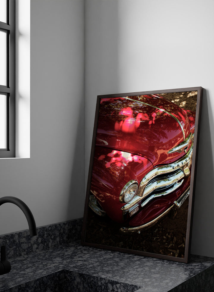 Close-up of a vintage red Chevrolet car body with chrome grille and reflections of trees on the glossy paint, photographed outdoors on mulch. Vintage Red Car by Shot by Clint automotive photography print featuring a classic Chevrolet, deep red paint, polished chrome, and sculptural vintage details.