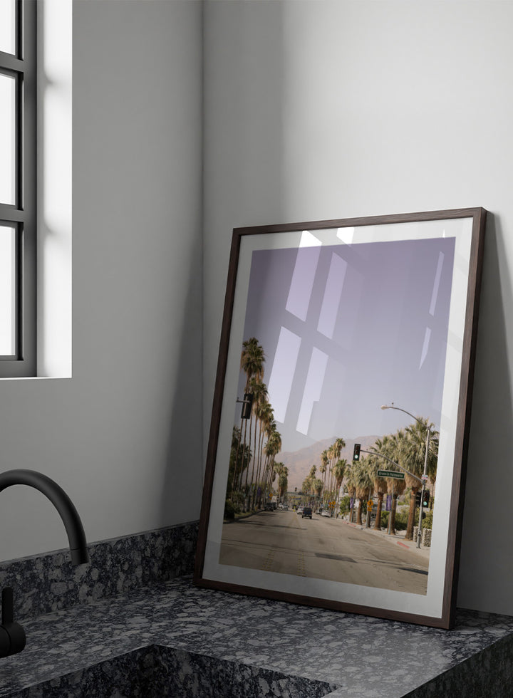 A framed art print featuring a scenic photograph of a palm-lined street in Palm Springs with mountains in the background. Palm-Lined Avenue by Shot by Clint travel photography print featuring a California road, tall palm trees, desert mountains, and soft sun-faded tones.