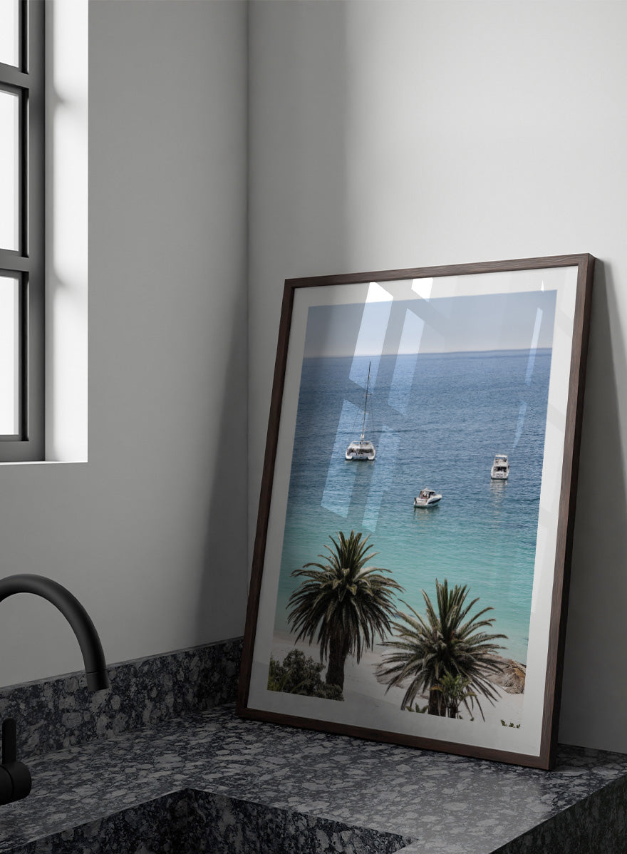 A framed coastal photograph showing a calm blue ocean with three boats, turquoise water near a white sandy shore, and two palm trees in the foreground. Tropical Serenity by Shot by Clint coastal photography print featuring turquoise water, sailboats, palm trees, and a bright tropical shoreline.