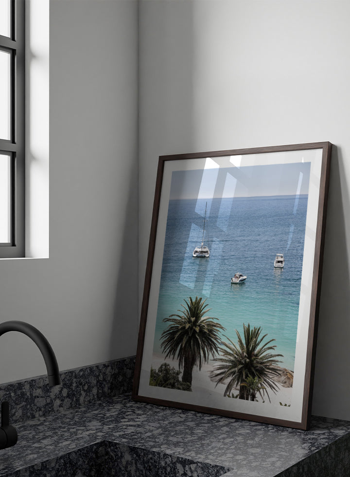 A framed coastal photograph showing a calm blue ocean with three boats, turquoise water near a white sandy shore, and two palm trees in the foreground. Tropical Serenity by Shot by Clint coastal photography print featuring turquoise water, sailboats, palm trees, and a bright tropical shoreline.