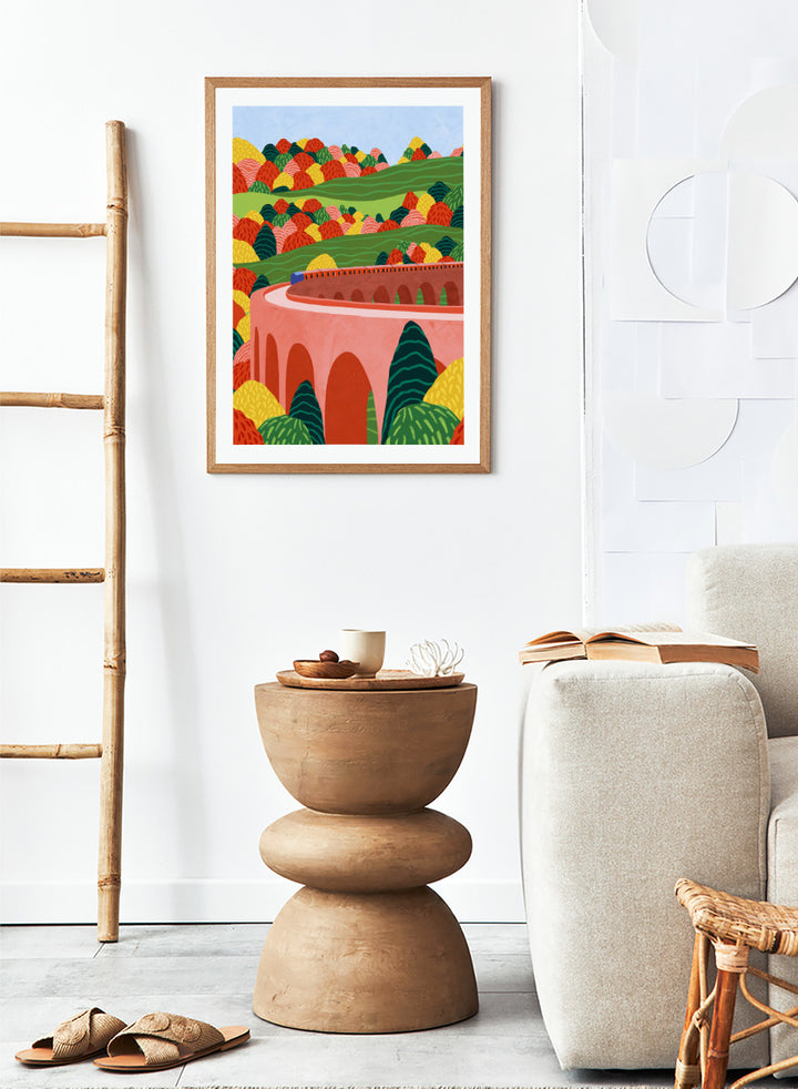 A framed art print featuring a stylized landscape illustration with rolling green hills, colorful patterned trees, and a pink arch bridge under a light blue sky. Winding Autumn Viaduct by Ceyda Alasar travel art print featuring a colorful train, arched bridge, rolling hills, and bold autumn foliage.